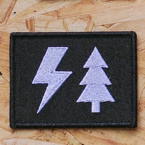 May include: A rectangular black embroidered patch featuring a light blue lightning bolt symbol on the left and a light blue stylized evergreen tree symbol on the right. The patch has a stitched border and a flat, textured fabric surface.