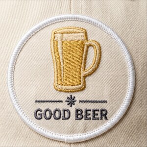May include: Embroidered patch featuring a golden beer mug with a frothy top, centered on a beige circle. The words "GOOD BEER" are embroidered below the mug in dark gray, with a decorative line and asterisk. The patch is bordered with white stitching.