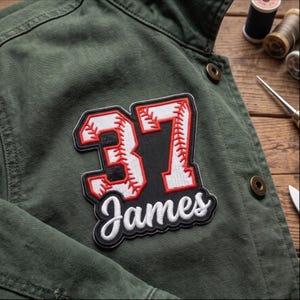 May include: An olive green jacket featuring a large embroidered patch with the number 37 in white with red baseball-style stitching and a black outline. Below the number, the name James is embroidered in white cursive script on a black background.
