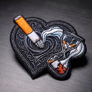May include: An embroidered patch shaped like a heart with a black background and intricate dark gray knotwork. The design features a lit cigarette with orange and white colors, and an hourglass containing cigarette butts and white smoke.