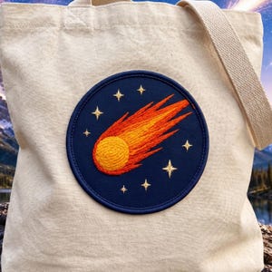 May include: A beige canvas tote bag featuring a circular dark blue patch centered on the front. The patch displays an embroidered orange and yellow comet with a textured, swirling head and a long tail, surrounded by small, stitched gold stars.