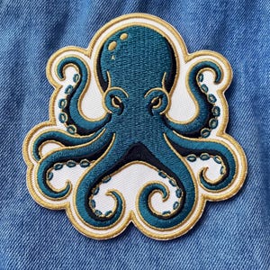 May include: An embroidered patch featuring a teal octopus with black accents and gold thread detailing. The octopus is centered on a white background with a gold border, showcasing eight tentacles with visible suction cups and a rounded head.