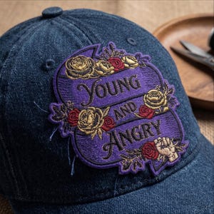May include: A dark blue denim baseball cap featuring a large, purple, embroidered patch. The patch displays the text "YOUNG AND ANGRY" in black lettering, surrounded by detailed gold and red floral accents and a small, raised fist icon.