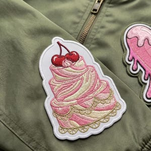 May include: Embroidered patch featuring a three-tiered pink cake with white frosting and red cherries on top. The patch has a white border and gold trim. The patch is on an olive green jacket.