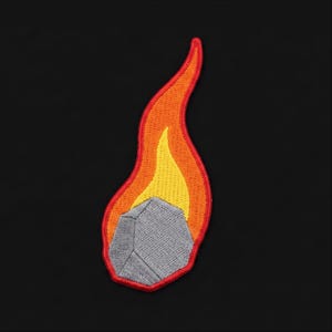 May include: An embroidered patch featuring a gray, faceted rock shape trailing a flame design. The flame consists of a bright yellow center surrounded by an orange layer, all outlined with a thick red border against a solid black background.