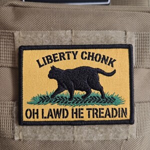 May include: A rectangular embroidered patch with a yellow background and a black border. It features a black cat silhouette walking on green grass. The text LIBERTY CHONK is above the cat, and OH LAWD HE TREADIN is below the cat in black.