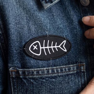 May include: An oval-shaped black embroidered patch featuring a white fish skeleton design with an X for an eye. The patch is stitched onto a dark blue denim jacket with a visible pocket seam, showcasing a clean, high-contrast graphic aesthetic.