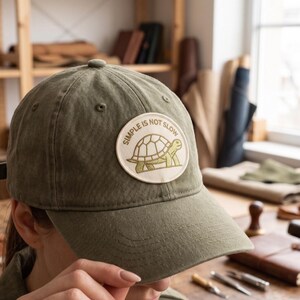 May include: An olive green baseball cap featuring a circular patch on the front. The patch displays a line-art illustration of a turtle and the text "SIMPLE IS NOT SLOW" in a circular arrangement. The cap has a structured crown and a curved brim.