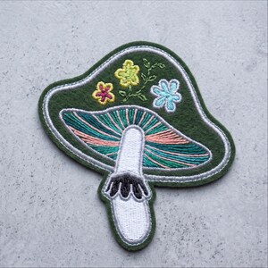 May include: An embroidered patch shaped like a mushroom on a dark green felt base. The mushroom cap features teal and pink radiating lines, topped with small yellow and light blue flowers. The stem is white with a black fringed ring detail.