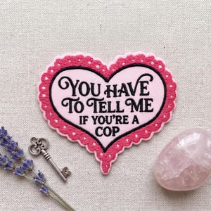 May include: A heart-shaped embroidered patch with a light pink center and a dark pink scalloped border. The patch features black embroidered text that reads: YOU HAVE TO TELL ME IF YOU'RE A COP. The design is centered on a textured fabric background.