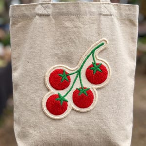 May include: A beige canvas tote bag featuring a central embroidered patch of a vine with four bright red tomatoes and green stems. The embroidery is outlined in a cream-colored border, creating a distinct graphic design on the fabric surface.