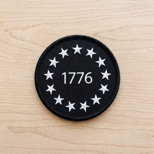 May include: A round black patch with white embroidered stars encircling the year "1776". The patch is on a light brown wooden surface.