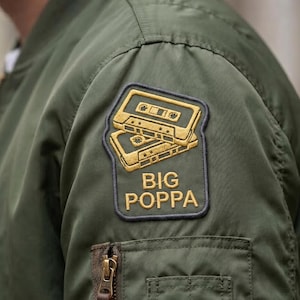 May include: Olive green bomber jacket with a cassette tape patch. The patch features two gold cassette tapes stacked on top of each other with the words "BIG POPPA" in gold lettering. The jacket has a zipper and a pocket.