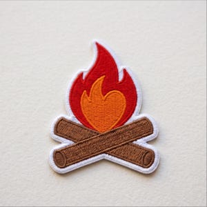 May include: An embroidered patch featuring a campfire design with two crossed brown logs at the base, topped with a bright orange inner flame and a larger red outer flame, all outlined with a thick white border against a plain background.