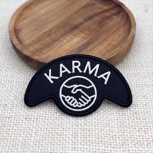 May include: A black embroidered patch with a crescent shape featuring the word KARMA in white capital letters above a circular icon of two hands shaking. The design is rendered in white thread against a solid black fabric background.