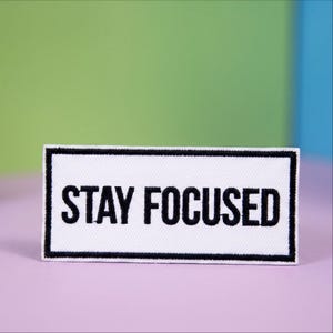 May include: A rectangular embroidered patch featuring a white background with a thick black border. The text STAY FOCUSED is stitched in bold black capital letters across the center of the patch, creating a clear and high-contrast design.