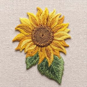 May include: A sunflower embroidery patch featuring vibrant yellow and golden petals, a textured brown center, and two green leaves at the base. The design is stitched with detailed thread work on a neutral, light-colored woven fabric background.