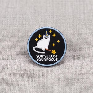 May include: A circular embroidered patch with a black background featuring a white cat wearing a blue wizard hat and holding a brown wand with a yellow star. Yellow stars surround the cat, and the text reads YOU'VE LOST YOUR FOCUS.