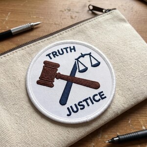 May include: A round white patch with the words "TRUTH" and "JUSTICE" in navy blue. The patch features a brown gavel and a navy blue scale. The patch is on a beige canvas pouch with a zipper.