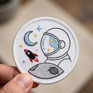 May include: Embroidered patch featuring an astronaut in a spacesuit, a crescent moon, stars, a rocket, and a planet. The patch is round with a white background and a stitched border. The astronaut's helmet has a blue and yellow design.