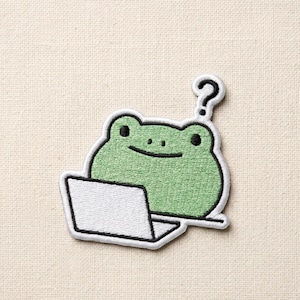 May include: Embroidered patch featuring a green frog with black eyes and a smile, sitting in front of a white laptop. A black question mark is above the frog's head. The patch has a white border and is on a beige background.
