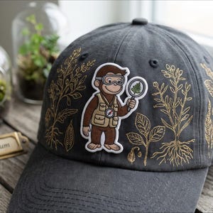 May include: A charcoal gray baseball cap featuring a central embroidered patch of a brown monkey wearing a tan vest, goggles, and holding a magnifying glass. The cap is decorated with gold-colored line art illustrations of various plants.