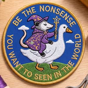 May include: A circular embroidered patch with a blue background and gold border. It features a white goose carrying an opossum wearing a purple wizard hat and robe with celestial patterns. The text reads: BE THE NONSENSE YOU WANT TO SEEN IN THE WORLD.