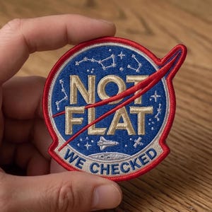 May include: A circular embroidered patch featuring a blue background with white constellations and stars. The text NOT FLAT is in gold, with a red swoosh across it. Below, the text WE CHECKED is in blue on a white arc, with a space shuttle icon.
