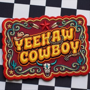 May include: A rectangular embroidered patch with a scalloped red border on a maroon background. It features the yellow text YEEHAW COWBOY, a brown cowboy hat, a lasso, a red star, two feathers, a bull skull, two cacti, and small gold stars.