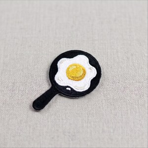 May include: An embroidered patch shaped like a black frying pan containing a sunny-side up egg with a bright yellow yolk and white egg whites. The patch features detailed stitching throughout, creating a textured appearance of a cooked breakfast.