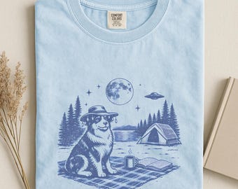May include: Light blue t-shirt with a graphic of a dog wearing a hat, camping under a full moon. The dog is sitting on a picnic blanket with a cup and a book. The shirt has the word "CHAMBRAY" printed on it.