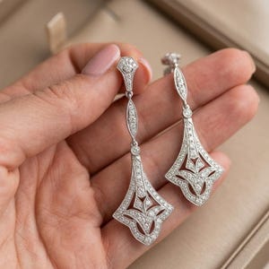 May include: A pair of Art Deco earrings. Each earring has a diamond-set design with a long, slender top and a geometric, fan-shaped bottom. The earrings are silver-toned and sparkling, with a vintage aesthetic.