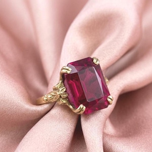 May include: A gold ring featuring a large, rectangular, deep red gemstone. The ring has an ornate, floral-inspired design around the gemstone and on the band. The ring is set against a soft, pink fabric background.