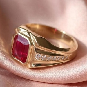 May include: A gold ring featuring a large, rectangular, deep red gemstone set in the center. The band is crafted from polished gold and includes a row of small, clear, round-cut stones embedded along the side of the shank, adding detail.