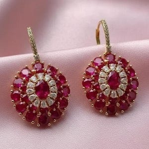 May include: A pair of gold-toned earrings with a floral design. Each earring features a central oval ruby surrounded by a halo of small diamonds and ruby-colored stones. The earrings are set against a pink background.