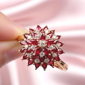 May include: A gold ring featuring a floral design. The ring's centerpiece is composed of numerous red gemstones and smaller white gemstones, creating a starburst effect. The ring is held against a soft pink background.