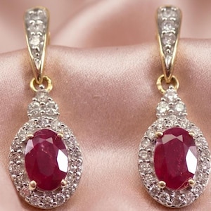Puede incluir: Un par de pendientes colgantes dorados. Cada pendiente presenta un gran rubí de corte ovalado rodeado de pequeños diamantes brillantes. La parte superior de los pendientes también tiene un diseño con diamantes.