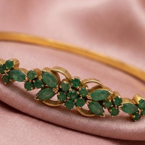 Puede incluir: Un brazalete dorado con un diseño floral. El brazalete presenta piedras preciosas verdes esmeralda con corte marquesa. La joya está sobre un fondo de satén rosa.