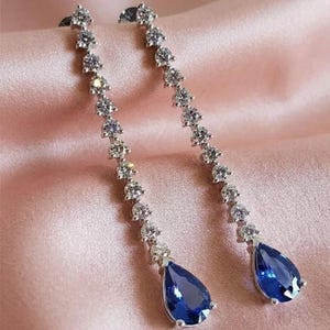 Puede incluir: Un par de elegantes pendientes colgantes. Cada pendiente presenta una serie de pequeñas piedras claras y brillantes que conducen a una gran gema azul zafiro en forma de pera. Los pendientes se colocan sobre un fondo rosa suave.
