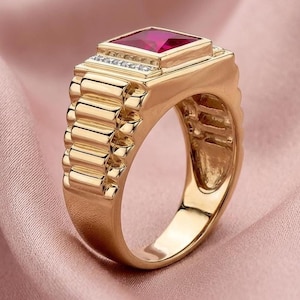 May include: A gold ring featuring a square-cut vibrant red gemstone set in the center. The band has a textured, link-style design on the sides, and a row of small, clear, sparkling stones is embedded along the base of the central square setting.