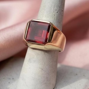 Puede incluir: Un anillo de sello de oro con una gran piedra preciosa de color rojo intenso y corte cuadrado engastada en el centro. La banda es lisa y pulida, con una construcción gruesa y sólida que se estrecha ligeramente hacia atrás, de diseño clásico.