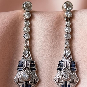 May include: A pair of drop earrings with a silver-tone metal setting. Each earring has a series of round, sparkling stones leading to a geometric design with dark blue accents and a larger central stone. The earrings are shown on a pink surface.