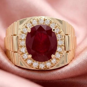 May include: A gold ring with a large, oval-cut ruby center stone. The ruby is surrounded by a halo of small, clear gemstones. The ring has a textured band.