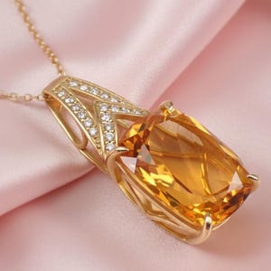 Könnte beinhalten: Eine goldene Anhänger Halskette mit einem großen, rechteckigen, bernsteinfarbenen Edelstein. Der Anhänger ist in einem goldenen Rahmen mit kleinen, klaren Akzentsteinen gefasst. Die Halskette ist ebenfalls aus Gold, vor einem weichen rosa Stoff.