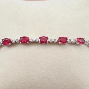 May include: A silver bracelet featuring oval-cut, vibrant red gemstones alternating with small, round, clear gemstones. The bracelet is displayed on a soft, off-white surface, showcasing its elegant design.