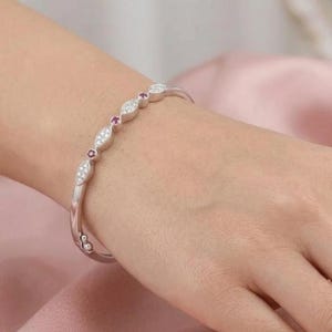 May include: A silver-toned bangle bracelet featuring a series of marquise-shaped links encrusted with clear white stones, alternating with small circular links set with deep red stones, secured by a hinged clasp mechanism on the side.