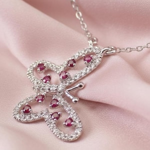 May include: A silver butterfly pendant necklace with a delicate chain. The butterfly is encrusted with small, clear crystals and features several red gemstones. The necklace is displayed on a pink satin background.