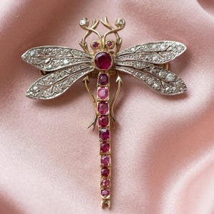 Puede incluir: Broche de libélula de oro y plata con detalles de rubíes y diamantes. El cuerpo de la libélula es una línea de rubíes, con una gran cabeza de rubí. Las alas están incrustadas con diamantes y las antenas tienen pequeños detalles de diamantes.