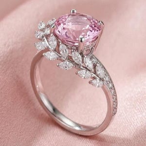 May include: A silver-toned ring featuring a large, round, faceted pink center stone held by four prongs. The band is adorned with a series of small, marquise-cut clear stones arranged in a leaf-like pattern extending from the central setting.