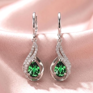 Pode incluir: Um par de brincos pendurados prateados com uma pedra preciosa verde vibrante em forma de gota no centro. Os brincos são adornados com pequenas pedras claras brilhantes e apresentam fecho de pressão. São exibidos sobre uma superfície de cetim rosa.